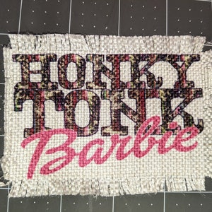 Może przedstawiać: Beige'owy łatek z burlapu z postrzępionymi krawędziami, na kt&oacute;rym widnieje napis "Honky Tonk Barbie" czarnymi i r&oacute;żowymi literami. Tekst "Honky Tonk" jest nadrukowany w czarno-brązowym wzorze lamparta.