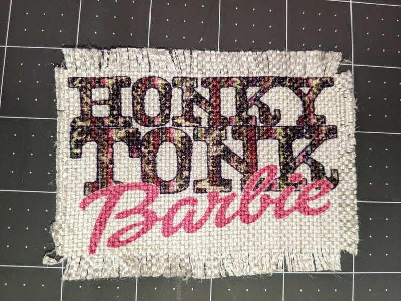 Może przedstawiać: Beige'owy łatek z burlapu z postrzępionymi krawędziami, na kt&oacute;rym widnieje napis "Honky Tonk Barbie" czarnymi i r&oacute;żowymi literami. Tekst "Honky Tonk" jest nadrukowany w czarno-brązowym wzorze lamparta.
