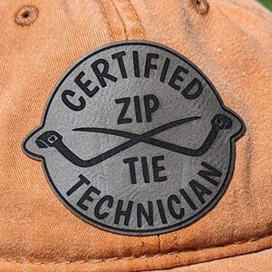 May include: A tan baseball cap with a gray patch. The patch reads "CERTIFIED ZIP TIE TECHNICIAN" in a circular design, with crossed zip ties in the center. The cap is made of a textured fabric.