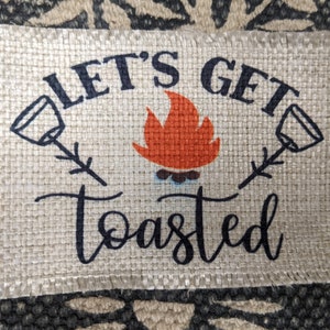 May include: A burlap fabric sign with the text "Let's Get Toasted" in black lettering. The sign features a campfire with orange flames and two black silhouettes of marshmallows on sticks.
