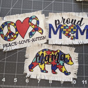 May include: Three white fabric patches with frayed edges. The first patch has a peace sign, a red heart, and a blue and yellow ribbon with the text "Peace. Love. Autism". The second patch has the text "Proud Mom" in blue with a blue and yellow puzzle piece heart. The third patch has a blue and yellow puzzle piece bear with the text "mama".