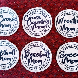 May include: Six round, white fabric patches with black text. Each patch has a different sport theme: Lacrosse, Cross Country, Wrestling, Softball, Baseball, and Soccer. Each patch says "Crazy Proud" and "Always Loud" around the sport name.