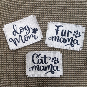 May include: Three white fabric patches with black text and paw prints. The patches read "dog mom", "fur mama", and "cat mama".