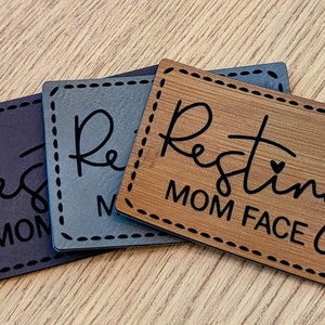 May include: Three wooden signs with a stitched border. The signs are different colors: black, gray, and brown. The signs all say "Resting Mom Face" with a small heart symbol.