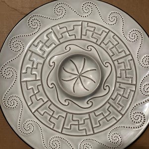 May include: White ceramic serving dish with a central dip bowl and decorative raised patterns. The dish has a black rim and a geometric design.