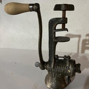 May include: Antique cast iron meat grinder with a wooden handle. The grinder has a screw clamp and a crank handle. The text "Everedy's No. 25" is visible on the side of the grinder.