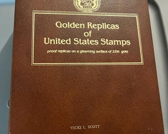 May include: A brown book titled "Golden Replicas of United States Stamps" with gold lettering and an eagle emblem. The text below reads "proof replicas on a gleaming surface of 22kt gold." The author's name, Vicki L. Scott, is at the bottom.