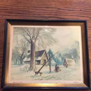 May include: A framed winter scene painting depicting a small wooden cabin with a snow-covered roof, a well, and two figures in the distance. The painting is set against a backdrop of trees and a snowy landscape. The text "Pilgrim Hill, Cape Cod" is printed in the bottom left corner.