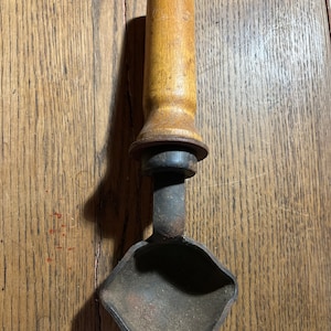 May include: A vintage wooden-handled metal ladle with a rounded bowl. The handle is made of wood and has a black metal ring at the top. The ladle is rusty and has a worn appearance.
