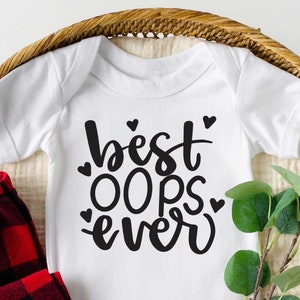 Könnte beinhalten: Ein weißer Baby-Body mit dem schwarzen Schriftzug "best OOps ever", verziert mit kleinen Herzen. Dazu eine rot-schwarz karierte Hose, braune Babyschuhe aus Leder und Grünzeug, alles in einem geflochtenen Korb.