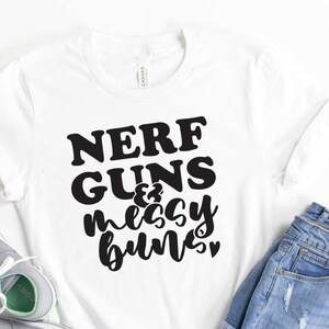 May include: A white t-shirt with black text that reads "NERF GUNS & messy buns". The shirt is laid out on a white surface with a pair of gray sneakers and a pair of blue denim shorts.