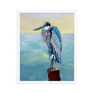 May include: A framed painting of a great blue heron standing on a dark red wooden post. The bird features shades of blue, gray, and white feathers with a long beak. The background depicts a calm body of water and a soft yellow sky.