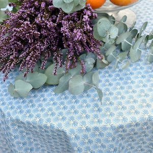 Peut inclure: Une nappe bleue et blanche avec un motif floral. La nappe est décorée d'un bouquet de fleurs violettes et de feuilles vertes.