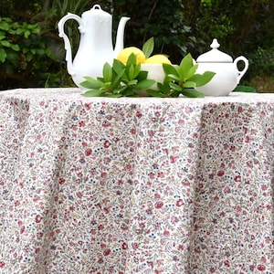 May include: A round tablecloth with a floral pattern in red, blue, and green on a cream background. A white teapot, sugar bowl, and creamer are on the table, along with lemons and greenery. The table is outdoors.