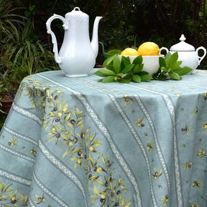 May include: A round table with a light blue tablecloth patterned with olive branches. A white porcelain coffee pot, a small sugar bowl, and a bowl of lemons are on the table. Green foliage adds a natural touch.
