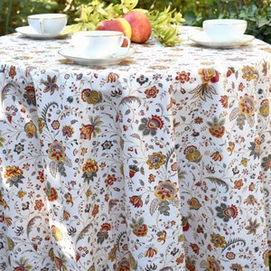 Peut inclure: Une nappe ronde blanche avec un motif floral dans les tons de rouge, jaune et vert. La nappe est posée sur une table avec trois tasses à thé et soucoupes et des pommes sur la table.