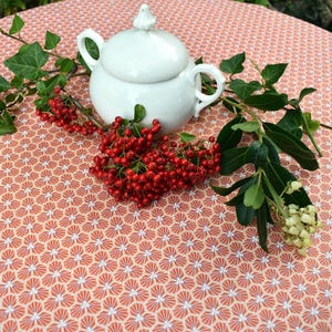 Può includere: Una teiera in ceramica bianca con coperchio e due manici è appoggiata su una tovaglia fantasia. Bacche rosse e fogliame verde sono disposti attorno alla teiera. La tovaglia ha un motivo floreale ripetuto in rosso e bianco.