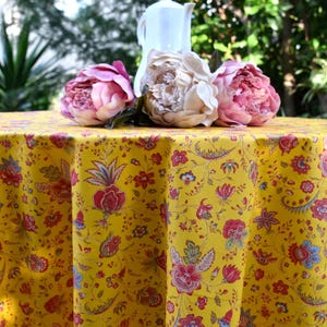Op de afbeelding: Een ronde gele tafelkleed met een bloemenpatroon, met rode, roze en blauwe bloemen. Drie grote pioenrozen en een witte kan liggen erop. Het tafelkleed staat buiten.