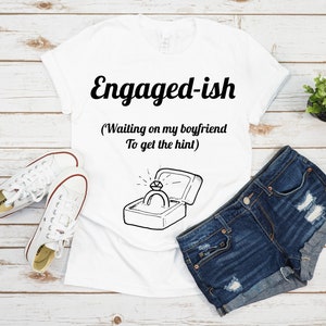 Könnte beinhalten: Weißes T-Shirt mit dem Text "Verlobtish (Warte darauf, dass mein Freund den Hinweis versteht)" und einer Zeichnung eines Rings in einer Schachtel.