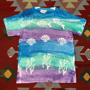 Puede incluir: Camiseta de manga corta con un diseño de acuarela que presenta esqueletos de peces, conchas marinas y coral. La camiseta tiene un esquema de color azul, morado y verde. La marca "Lady Rustle" está impresa en la etiqueta.