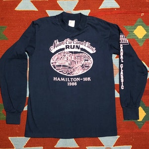 May include: A navy blue long-sleeve t-shirt with a pink graphic of a boat on a canal. The graphic says "Miami Erie Canal Classic Run" and "Hamilton-10K 1986". The right sleeve has the text "Miami Erie Canal Classic" printed vertically.