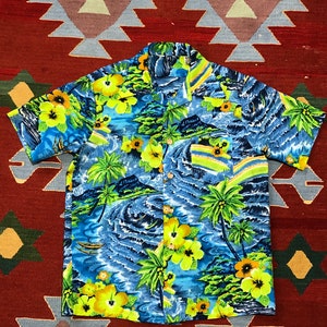 Op de afbeelding: Een shirt met korte mouwen en een tropische print. Het shirt heeft een blauwe achtergrond met gele hibiscusbloemen, palmbomen en oceaangolven. Het heeft een klassieke kraag en een enkele borstzak.