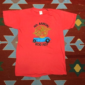 May include: A coral-coloured t-shirt with a graphic for the "4th Annual Eville Dron Rod Fest." The design features a blue car with yellow accents and text. The shirt is likely vintage and made of a soft material.