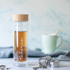 Op de afbeelding: Een glazen theefles met een bamboe deksel en een roestvrijstalen infuser. De fles is gevuld met bruine thee en heeft de naam "Rebecca" erop geschreven. De fles staat op een wit houten oppervlak met een grijze doek en losse theebladeren.