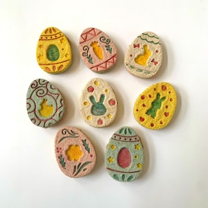 May include: Nine pastel-coloured Easter egg-shaped cookies with various designs, including bunnies, carrots, and flowers.