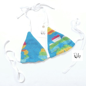 Puede incluir: Top de bikini azul con un diseño de paisaje colorido. Las copas triangulares están conectadas por correas blancas que se atan en el cuello y la espalda. Una etiqueta con la palabra "Ruby" está adjunta.