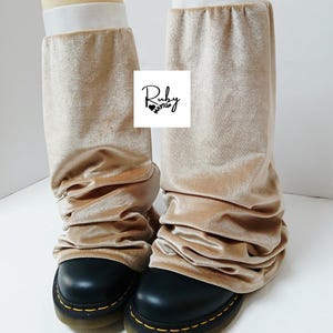 May include: A pair of tan velvet leg warmers with a white elastic band at the top, worn over black boots. The leg warmers are scrunched and gathered. The boots have yellow stitching around the soles. The Ruby Maytup logo is visible.