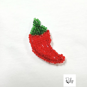 May include: A chili pepper patch crafted from small, vibrant red and green glass beads. The pepper shape is filled with translucent red beads, while the stem is composed of deep green beads. A small white logo with the text Ruby Maytup is visible.