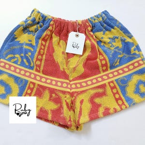 Pode incluir: Calções de tecido atoalhado com cintura elástica. Os calções apresentam um padrão vibrante em vermelho, amarelo e azul, com uma borda decorativa. Uma etiqueta com o logotipo Ruby Maytup está anexada.
