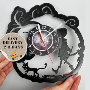 May include: Black vinyl record clock with a silhouette of a witch and a black cat. The clock face is white with black numbers and a black and white moon design. The clock hands are black and red.