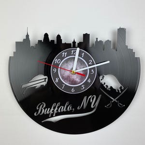 May include: Black vinyl record clock with a white clock face and black hands. The clock is shaped like the Buffalo, NY skyline with a buffalo silhouette and the text "Buffalo, NY".