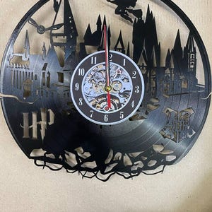 May include: Black vinyl record clock featuring a silhouette of Hogwarts castle and a wizard flying on a broomstick. The clock face has Roman numerals and a white background. The clock is labeled "HP" in the lower left corner.