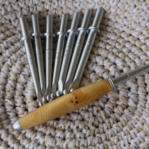 May include: A collection of silver crochet hooks with wooden handles, displayed on a beige and white crocheted surface. The hooks are marked with sizes D/3, E/4, F/5, G/6, H/8, and I/9. One hook features a light-coloured wooden handle.