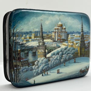 May include: A rectangular lacquered box featuring a detailed hand-painted winter scene of a city with snow-covered trees, a stone bridge, and a large cathedral with golden domes. The artwork depicts figures walking along a snowy path in blue tones.