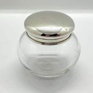 May include: A small, clear glass jar with a rounded, bulbous body and a smooth, polished silver-toned metal lid. The container features a wide opening and a simple, clean design, showcasing a transparent glass base topped with a reflective metallic cap.