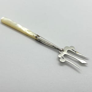 May include: A silver serving fork featuring a three-pronged head with decorative scalloped edges and a smooth, elongated handle made of iridescent white mother-of-pearl. The metal neck connects the handle to the tines, which bear small hallmarks.