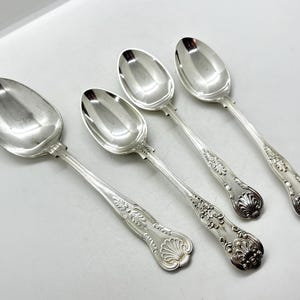 May include: Four silver spoons with decorative handles. The spoons have oval bowls and ornate detailing on the handles, including floral and shell-like patterns. The spoons are arranged on a white surface.