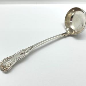 Könnte beinhalten: Eine silberne Schöpfkelle mit rundem Kessel und langem, gebogenem Griff. Der Griff weist verzierte Muschel- und Blumendesigns auf. Die Schöpfkelle besteht aus einem glänzenden, reflektierenden Metall, wahrscheinlich Silber oder einer silberfarbenen Legierung. Es ist ein Küchenutensil.