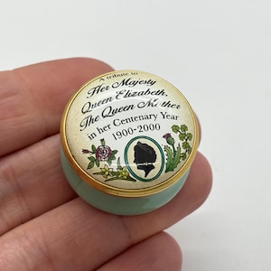 May include: A small, round, light blue trinket box with a gold rim. The lid features text that reads "A tribute to Her Majesty Queen Elizabeth, The Queen Mother in her Centenary Year 1900-2000." The lid also has a silhouette of a person.