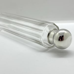 May include: A clear glass bottle with a long, cylindrical, fluted body and a smooth, rounded, polished silver-tone metal cap. The bottle features vertical ridges running along its length, ending in a circular neck topped by the reflective metal stopper.