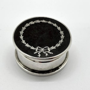 May include: A round silver-toned metal pill box featuring a black circular inlay on the lid. The lid is decorated with a silver-coloured engraved laurel wreath design that is tied with a bow at the bottom centre. The sides of the box are smooth.