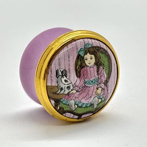 May include: A small, round, lavender trinket box with a gold-colored rim. The lid features a painted scene of a girl in a pink dress with a dog. The background is a pink and white striped pattern. The box is a decorative item.