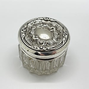 May include: A small, round antique jar with a silver lid and a clear, faceted glass base. The lid is intricately decorated with floral and scrollwork patterns. The jar is likely used for storing small items or cosmetics.