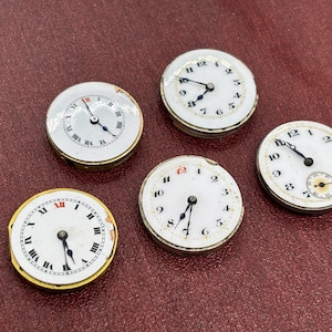 Könnte beinhalten: Fünf antike Taschenuhrzifferblätter mit weißen Zifferblättern, schwarzen Zeigern und römischen oder arabischen Ziffern. Die Zifferblätter sind rund, mit gold- oder silberfarbenen Rändern und liegen auf einer weinroten Oberfläche.