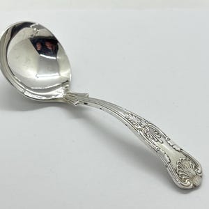 Könnte beinhalten: Eine silberne Schöpfkelle mit rundem Napf und dekorativem Griff. Der Griff weist ein verziertes, graviertes Design mit floralen und scrollartigen Motiven auf. Die Schöpfkelle besteht aus einem glänzenden, reflektierenden Metall, wahrscheinlich Silber oder einem versilberten Material.