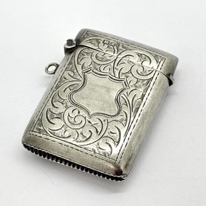 May include: A silver rectangular match safe with ornate, engraved floral and scrollwork designs. The hinged lid has a small loop for a chain. The body features a central blank cartouche. The edges are finely detailed.